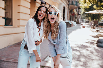 Amazing long-haired blonde girl fooling around on the street with friend. Photo of two carefree ladies chilling outdoor together.