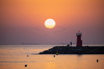 Sunrise at Guardamar jetty in Spain. A cyclist looks at the sea from the lighthouse with the island of Tabarca in the background. A flock of birds in the sky.