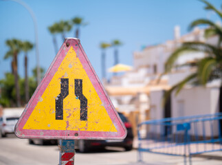 A yellow red round road sign on a small street in Spain. The roadway narrows at the construction site. The background is out of focus with nice bokeh.