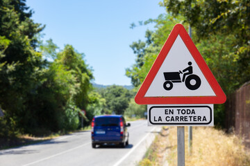 A triangular red and white warning sign on a country road in summer. It warns of a tractor. Translation: Applies to the entire street.
