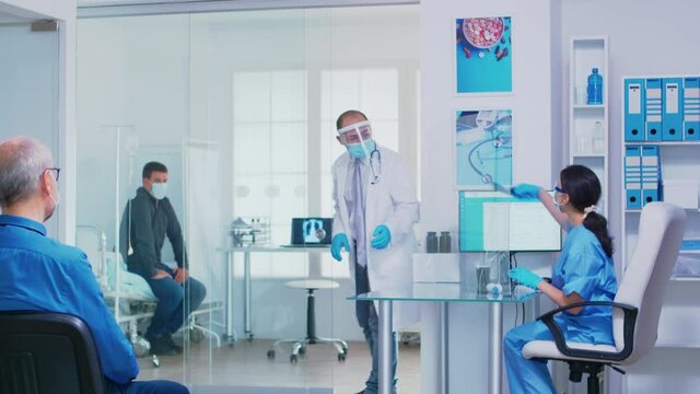 Nurse giving doctor patient radiography wearing face mask against coronavirus. Assistant with visor pusing disabled senior woman in wheelchair through waiting area.