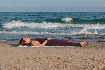 Young woman meditating while lying down on a yoga mat at the beach. Natural morning light shot with the Mediterranean Sea in the background.