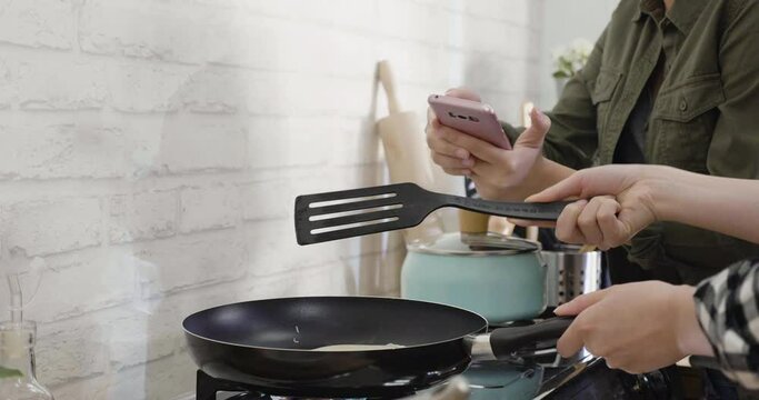 close up view of two female hands doing their stuff in modern kitchen. one girl is cooking crepe and turning side on hot pan by frying spoon. friend taking picture recording process on smart phone.