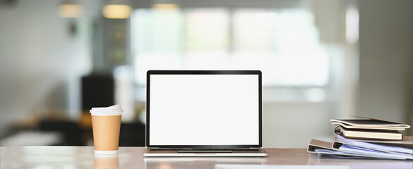 A white blank screen computer laptop is putting on a white table surrounded by a coffee cup and stack of books.