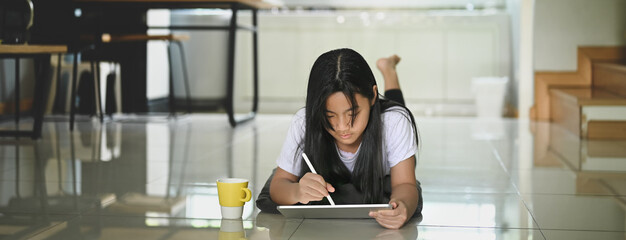 A little girl is using a computer tablet and stylus pen while lying on the living room ground floor.