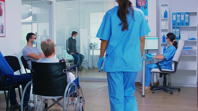 Doctor with face mask against covid19 discussing with nurse in hospital waiting area. Disabled senior woman in wheelchair waiting for examination. Assistant working on reception computer.