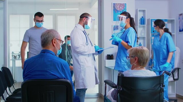 Medical team with face mask against coronavirus discussing in hospital waiting room about patient diagnosis. Senior man talking with disabled woman in wheelchair. Patient waiting for examination.