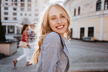 Emotional blonde woman posing with happy smile on the street. Debonair caucasian girl enjoying photoshoot on city background. - obrazy, fototapety, plakaty