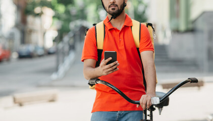 Online map application for courier for work. Serious guy with beard with backpack, looks at smartphone, near bicycle