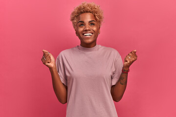 Portrait indoors of a dark-skinned girl with a short haircut who finally got what she really wanted. Girl in casual clothes stands on a background of a pink wall and is sincerely happy.