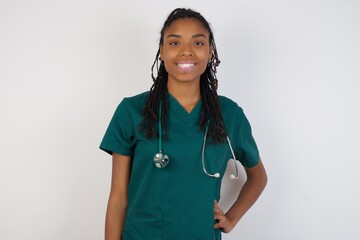 Studio shot of cheerful doctor woman keeps hand on hip, smiles broadly, anticipates for surprise prepared by friends on her birthday, isolated over gray background.