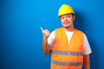 Young fat asian man over blue background wearing contractor uniform and safety helmet pointing and showing with his finger