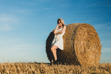 Naklejka na ściany i meble blonde girl is staying by a rolled haystack in field in sunset time