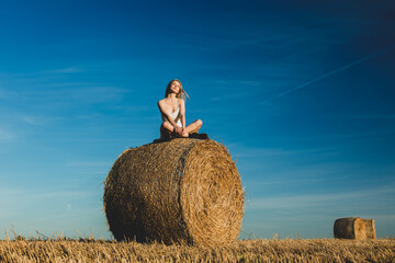Naklejka na ściany i meble blonde girl is sitting on rolled haystack in field in sunset time