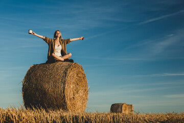 Naklejka na ściany i meble blonde girl is sitting on rolled haystack with cup of coffee in field in sunset time