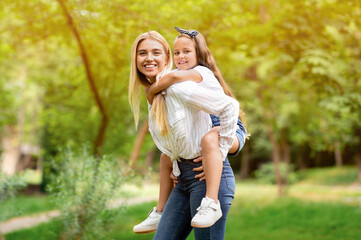 Fototapeta premium Joyful Mom Carrying Little Daughter Piggyback Having Fun In Park