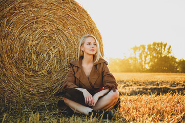 Naklejka na ściany i meble blonde girl is sitting by a rolled haystack in field in sunset time