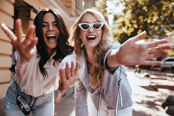 Blonde woman in elegant sunglasses posing with friend. Positive stylish girls smiling on city...