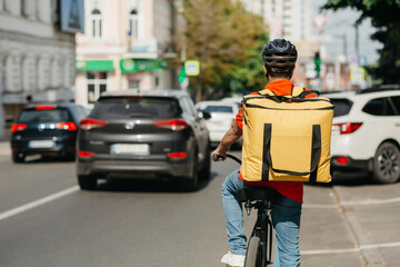 Male courier with helmet and big yellow backpack ride on bicycle along road on street
