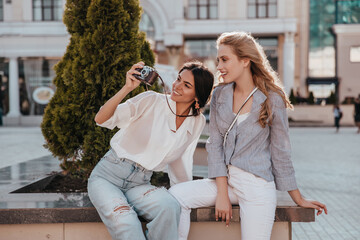 Fototapeta premium Female photographer in white blouse spending time with friend. Attractive brunette woman with camera taking pictures.
