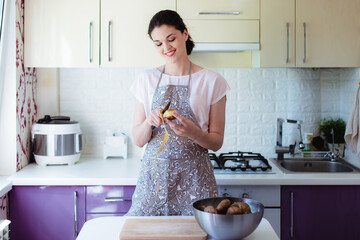 Young woman in kitchen in apron peeling potatoes