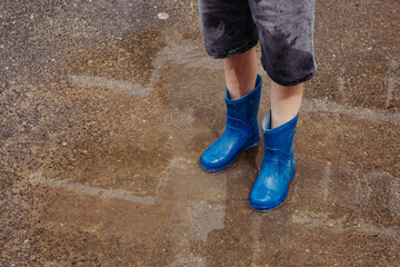 Children in the summer after the rain. Close-up of a boy's feet in blue rubber boots standing in a puddle on a walk. Children walk in the open air after the rain. Waterproof shoes for boys.