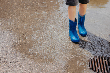 Children in the summer after the rain. Close-up of a boy's feet in blue rubber boots standing in a puddle on a walk. Children walk in the open air after the rain. Waterproof shoes for boys.