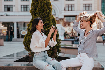 Beautiful female photographer taking picture of her funny sister. Excited blonde girl posing in front of camera.
