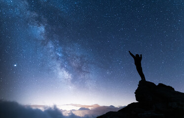 Fototapeta premium Person with arms in the air contemplating the vastness of the universe. Silhouette of a man under the milky way and starry magical sky. Real outdoor adventure and wild life concept.