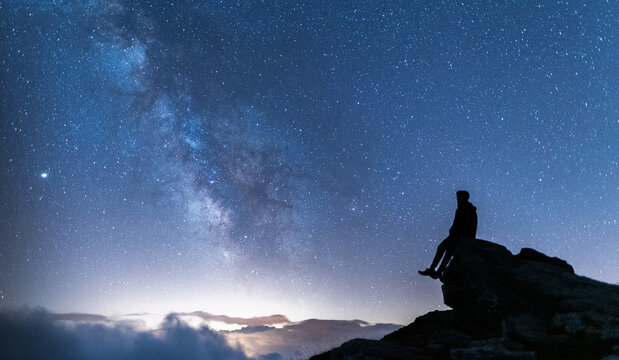 Person sitting on a rock contemplating the vastness of the universe. Silhouette of a man under the Milky Way and the magical starry sky. Real outdoor adventure and wildlife concept.