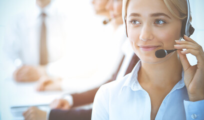 Cheerful smiling business woman with headphones consulting clients. Group of diverse phone operators at work in sunny office.Call center and business people concept