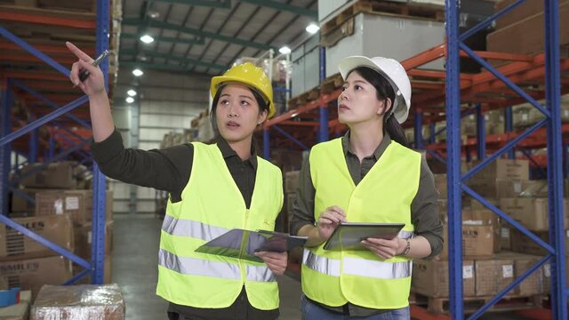 two female team partners meeting at warehouse and discussing about corporate work. beautiful lady coworkers point finger around in stockroom and talking. women in hard hat and safety vest working