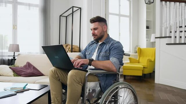 Likable concentrated high-skilled modern man with beard sitting in wheelchair and remotely working at home on laptop