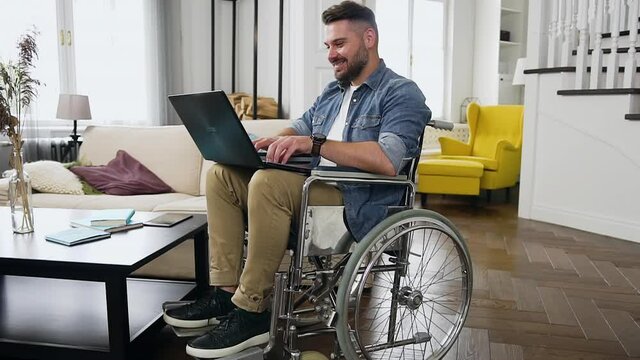 Handsome smiling satisfied young bearded man in wheelchair remotely working on personal computer at home