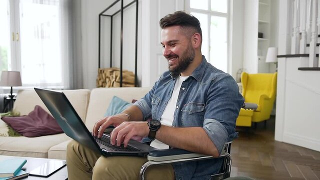Close up of good-looking modern joyful bearded young guy in casual clothes which sitting in wheelchair at home and remotely working on laptop
