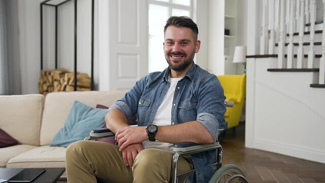 Handsome happy cheerful young bearded guy in wheelchair looking at camera with lucky smile on the home interier background