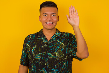 Young handsome man standing over yellow isolated background Waiving saying hello or goodbye happy and smiling, friendly welcome gesture © Jihan
