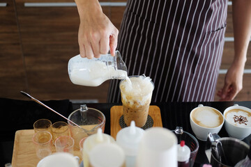 Asian barista young man pouring fresh milk in a plastic glass of latte iced coffee for according to order at counter bar in the morning at modern cafe.