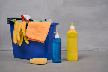 Close up shot of plastic bucket with cleaning supplies and bottles with detergents standing on the floor against grey wall