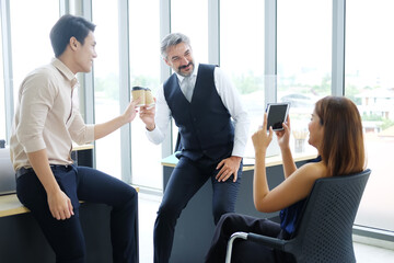 Smiling young Asian and Senior Caucasian businesspeople teamwork relaxing and holding a mug of hot coffee for take a photo with smartphone at modern office. Business and lifstyle concept