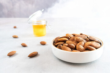 A full white bowl of peeled almonds sits on a light gray concrete surface. A small jar of honey stands in the background. Healthy food concept. May Help Lower Cholesterol Levels