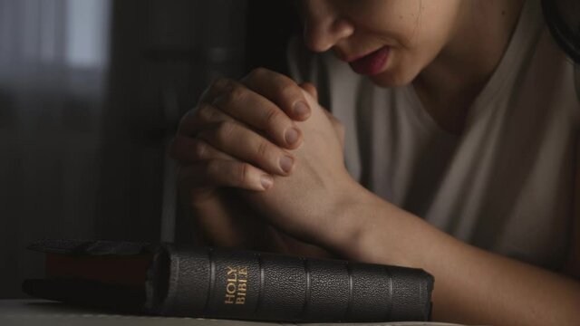 A woman prays with her hands crossed over a Bible for God's blessing in a dark room.