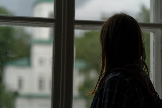 Close up of young woman looking out window in unlit room. Female silhouette observing through window.