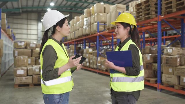 Happy female warehouse workers talk while preparing shipment for distribution in industrial warehouse. two chinese women employees having great deal in stockroom. ladies wear hard hat for protection