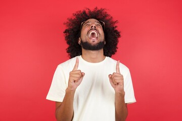 Charming carefree man with positive expression, points up with both index fingers, dressed in...