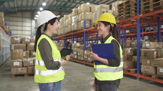 Two business women partners in helmets and safety vests shaking hands in warehouse. cheerful chinese ladies workers discussing laughing joyful. happy teammates handshaking before work in stockroom
