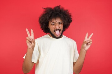 Indoor portrait of young caucasian male isolated on gray background with optimistic smile, showing peace or victory gesture with both hands, looking friendly. V sign.