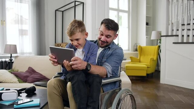Close up of likable positive contented happy family of son and his father in wheelchair which celebrating victory in video game on tablet pc