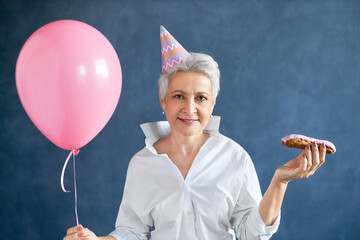 Horizontal image of stylish attractive middle aged businesswoman with pixie hairdo holding dessert and air balloon having fun at birthday party, celebrating 50th anniversary. People and aging concept