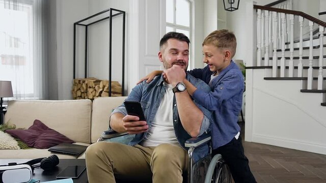 Joyful good-looking modern happy family of bearded dad in wheelchair and smiling son having fun together at home ,close up